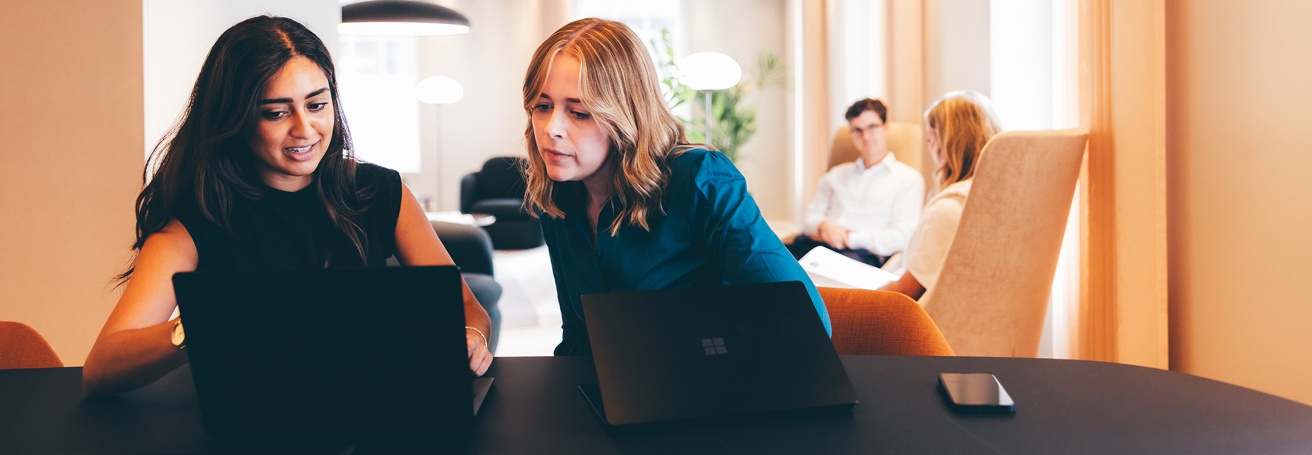 Two women work on laptops at a table, engaged in discussion. In the background, a man and woman converse on a sofa in a modern office setting.
