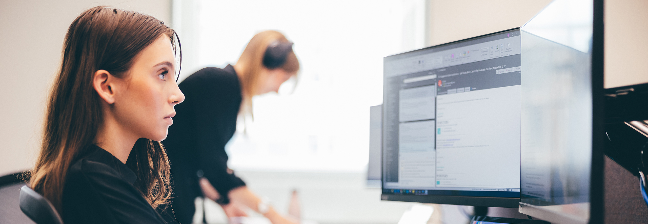 A woman intently looks at a computer screen displaying text, in an office with another blurred figure in the background working on a laptop.