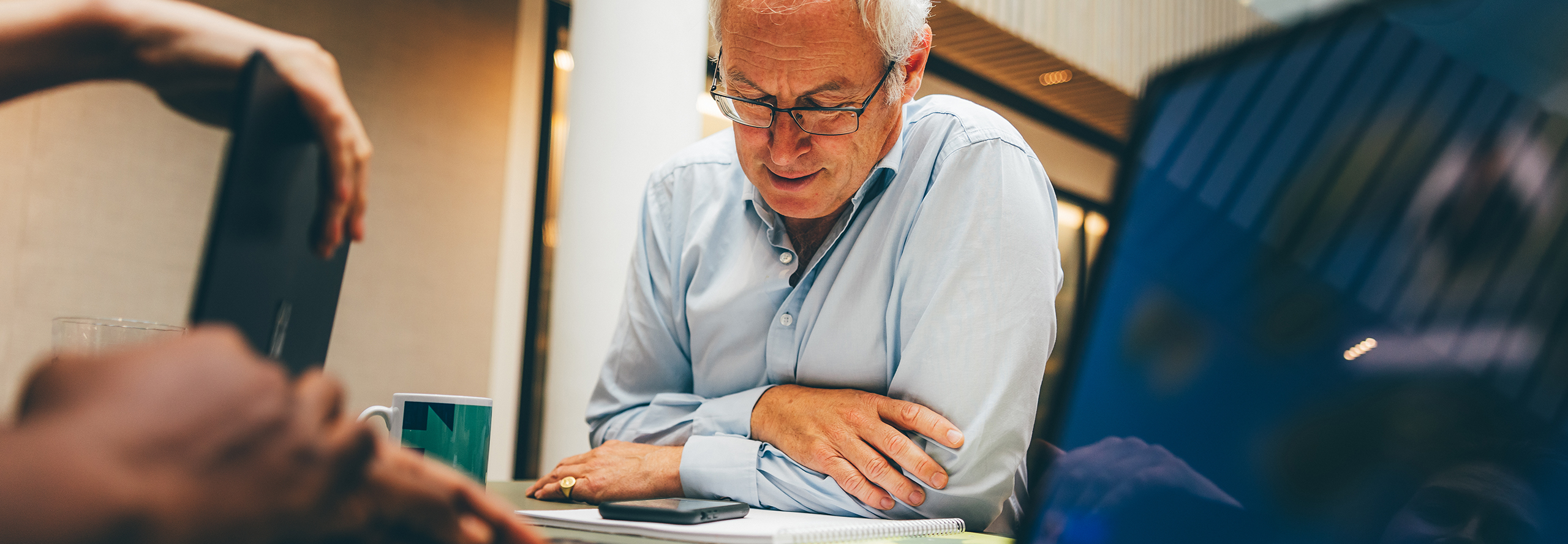An older man in glasses leans over a tablet, surrounded by a busy office environment, with a mug and notebook nearby.