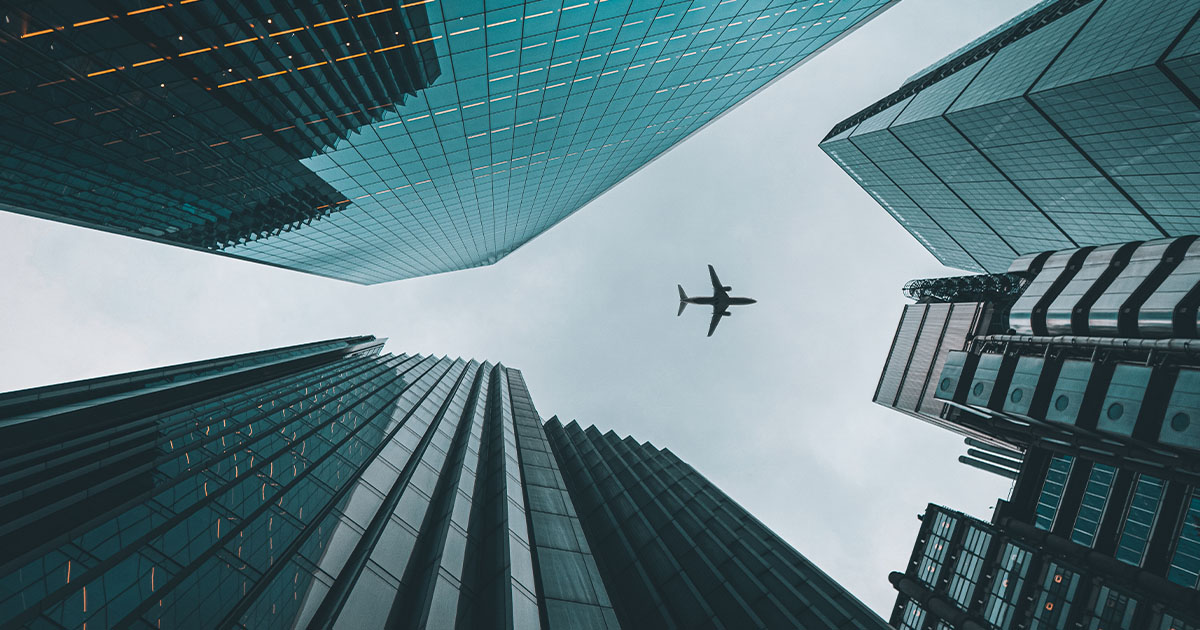 A plane flies overhead, seen from below, surrounded by towering glass skyscrapers in a cityscape, against a cloudy sky.