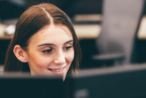 A person is smiling while looking at a computer screen, sitting in an office environment with blurred desks and chairs in the background.