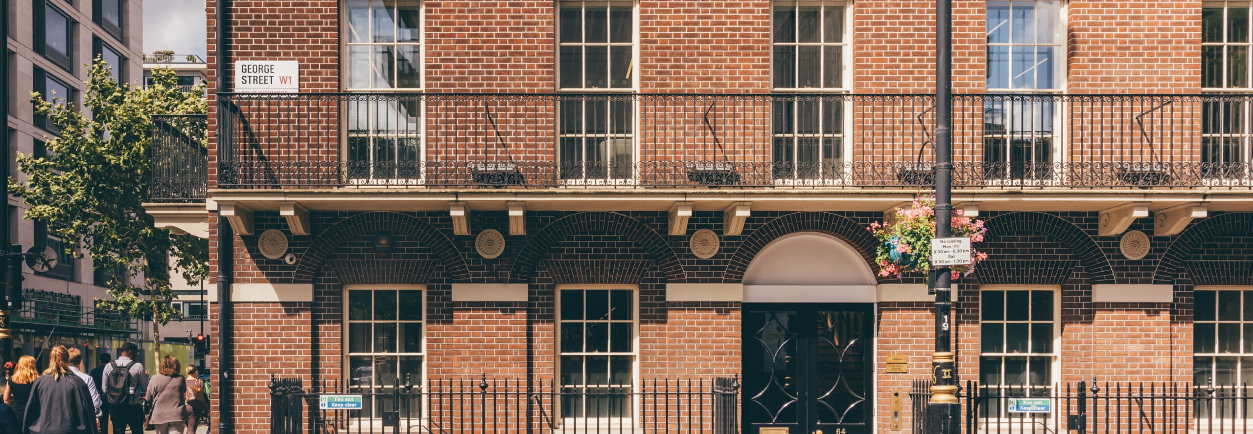 A red-brick building features black iron balconies and arched windows. Pedestrians walk nearby. Signs read "George Street W1" and "No loading Mon-Fri 8 am-9:30 am and Sat 8 am-1:30 pm". Another sign states "Fire exit Keep clear".